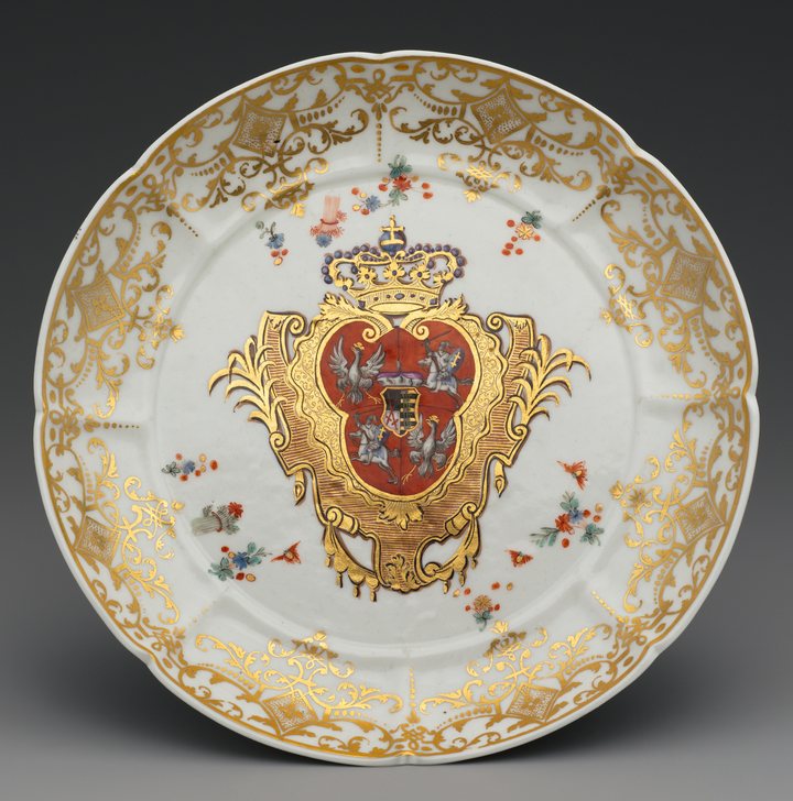 Prato de porcelana branca com borda recortada, ornamentos dourados e, no centro, um brasão vermelho e dourado sob uma coroa.