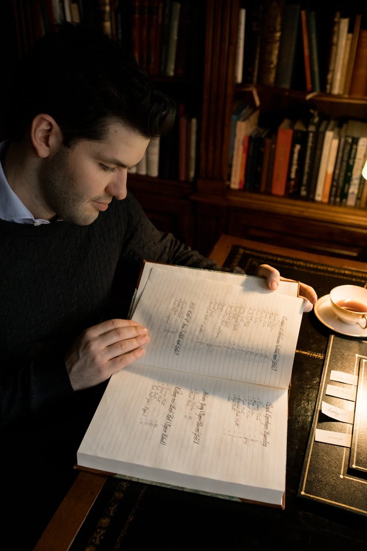 Rafa de Campos Salles lê um caderno manuscrito numa biblioteca de estantes escuras, com uma xícara de chá sobre a mesa.
